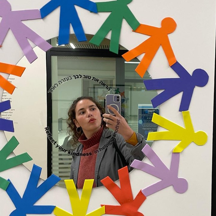 נגה מצלמת סלפי במראה שמוקפת בדמויות נייר צבעוניות שמחזיקות ידיים